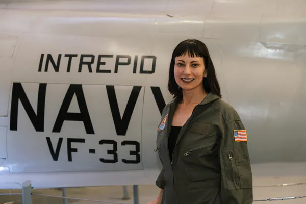 A guest in an olive flight suit smiles in front of a jet marked Intrepid Navy VF-33.